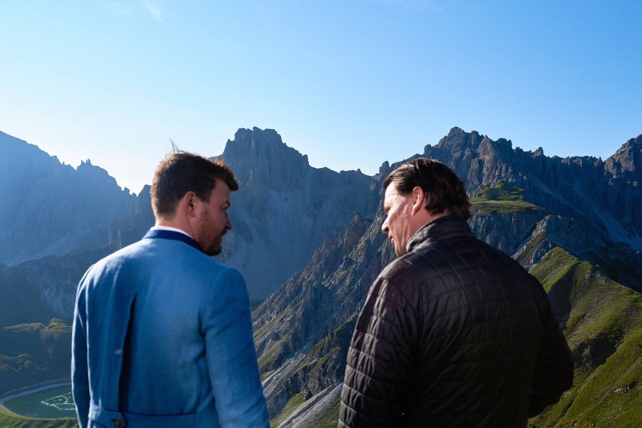 Viewed from the back, two men speak in front of a mountainous landscape.